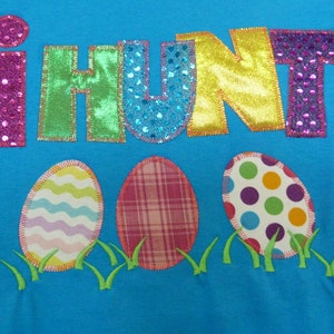 May include: A blue t-shirt with the words "i hunt" in colorful, glittery letters. Three embroidered Easter eggs are below the text, one with a plaid pattern, one with a wavy pattern, and one with polka dots.
