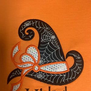 May include: An orange t-shirt featuring an embroidered black witch hat with a white spiderweb pattern. The hat is accented by a white ribbon with iridescent sequins and an orange border. The word Wicked is embroidered in black below the hat.