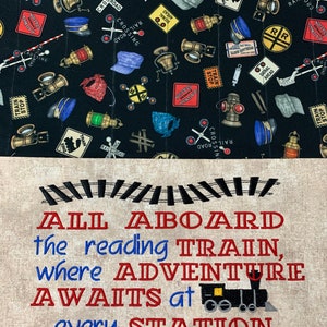 May include: A black fabric tote bag with a train theme. The bag has a quilted design and features a variety of train-related images, including train tracks, railroad crossing signs, and train engines. The bag has a white fabric bottom with the text "ALL ABOARD the reading TRAIN, where ADVENTURE AWAITS at every STATION" embroidered in red and blue.