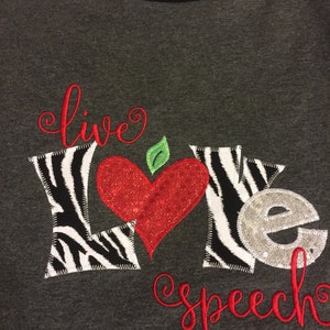 May include: A gray and black baseball style t-shirt with the words "live" and "speech" embroidered in red. The word "love" is embroidered in black and white zebra print with a red heart in the middle.