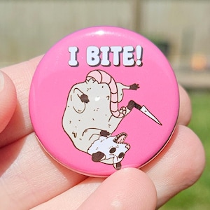 May include: A round, pink button with the text "I BITE!" in white. The button features a cartoon opossum holding a knife. The opossum is light beige with a white face and dark brown accents.
