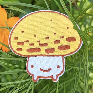 Puede incluir: Un hongo de fieltro amarillo con un tallo blanco y una cara sonriente. El hongo está bordado con detalles marrones.