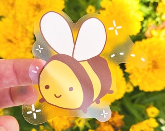 May include: A clear sticker with a cartoon bee design. The bee is yellow and brown with a smiling face and white wings. The sticker is surrounded by a white background with small white stars.
