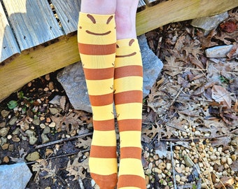 May include: A pair of yellow and brown striped knee-high socks with a smiling face design. The socks are worn on a person's legs.