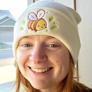 May include: Cream-colored beanie with an embroidered cartoon bee design. The bee is yellow and brown with white wings, surrounded by green leaves. The beanie is made of a soft, knitted material.