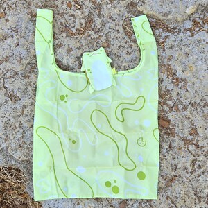 May include: A light green reusable shopping bag with a white and green abstract pattern. A small green frog-shaped pouch is attached to the bag.