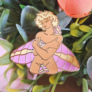 May include: A gold enamel pin depicting a nude figure with moth wings. The figure has blonde hair and is embracing itself. The wings are pink and yellow. A moth is on the figure's shoulder and another is on its lower body.