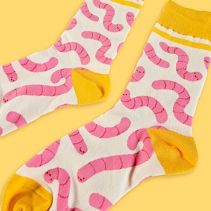 May include: A pair of white socks with a repeating pattern of pink cartoon worms. The socks have yellow heels and toes, and a yellow band at the top.