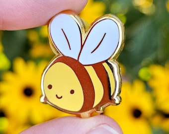 May include: A gold-colored enamel pin of a cartoon bee with a smiling face, brown stripes, and white wings.