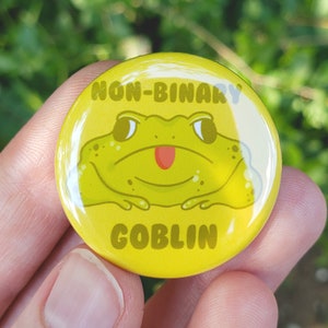 May include: Yellow button with a cartoon green frog with a red tongue and the text "Non-Binary Goblin" in black.