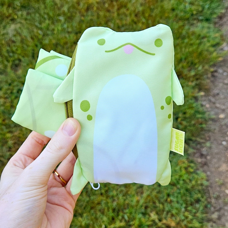 May include: A green frog-shaped reusable shopping bag with a white belly and a pink tongue. The bag is folded and held in a hand. The bag has a small tag that says "vegan" on it.