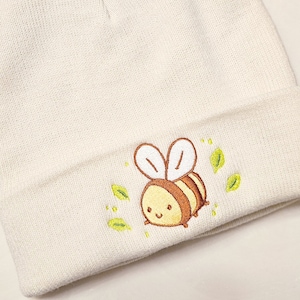 May include: Cream-colored beanie hat with an embroidered cartoon bee design. The bee is yellow and brown with white wings, surrounded by green leaves. The hat is made of a soft, knitted material.