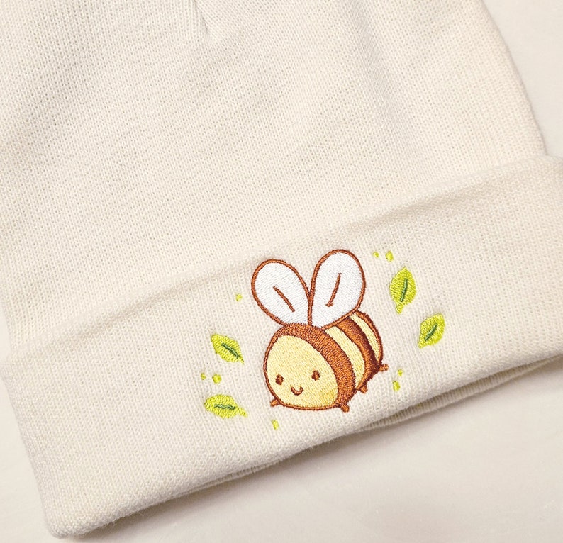 May include: Cream-colored beanie hat with an embroidered cartoon bee design. The bee is yellow and brown with white wings, surrounded by green leaves. The hat is made of a soft, knitted material.
