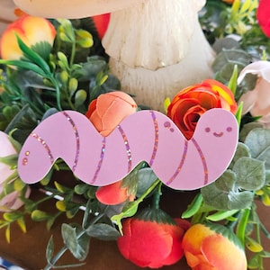 May include: A light pink, wavy worm-shaped sticker with glitter accents. The sticker has a smiling face and is surrounded by colorful flowers and greenery. The sticker is a whimsical and decorative item.