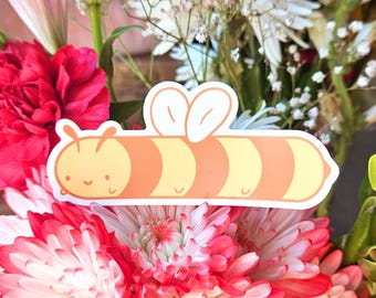 May include: A yellow and orange cartoon bee sticker with a smiling face and wings. The bee is on a white background and is surrounded by pink and white flowers.