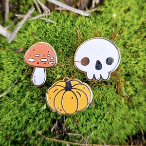 May include: Three enamel pins featuring a white skull, a red and white mushroom, and a yellow pumpkin. The pins are on a green mossy surface.