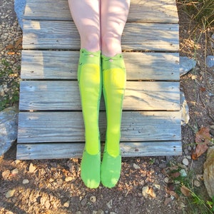 May include: A pair of green knee-high socks with a frog face design. The socks are worn by a person sitting on a wooden surface.