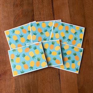 May include: A collection of square cards with a repeating pattern of yellow and orange pineapples on a light blue background. The cards have white borders and are arranged on a wooden surface.