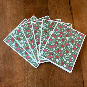 May include: A stack of greeting cards with a green background featuring a pattern of red ladybugs, white flowers, and small pink flowers. The cards have a white border and are arranged on a wooden surface.