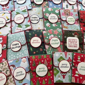 May include: A collection of 20 handmade Christmas gift card holders. The holders are made from patterned paper and feature various Christmas themes, including snowmen, Christmas trees, and ornaments. The holders are decorated with red, green, and white paper and have a small pocket for a gift card.