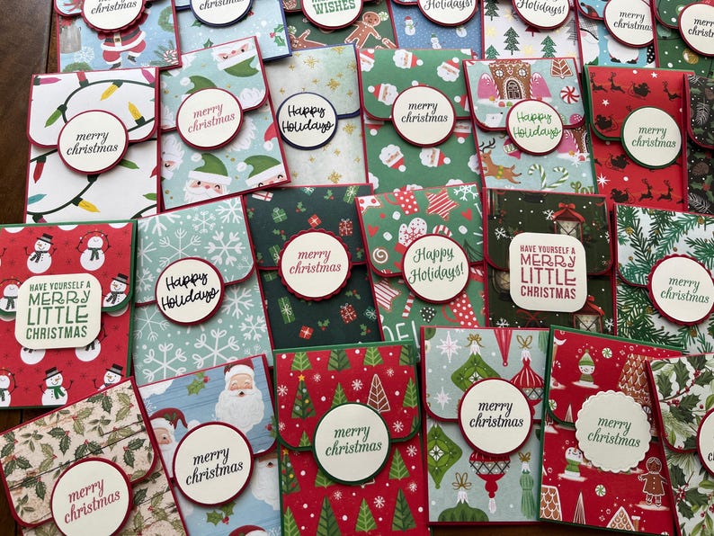 May include: A collection of 20 handmade Christmas gift card holders. The holders are made from patterned paper and feature various Christmas themes, including snowmen, Christmas trees, and ornaments. The holders are decorated with red, green, and white paper and have a small pocket for a gift card.