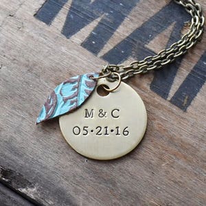 May include: A brass-colored pendant necklace with a chain. The pendant has a round tag with "M & C 05.21.16" stamped on it, and a leaf-shaped charm with blue and brown patterns. The necklace rests on a wooden surface.