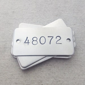 Op de afbeelding: Een stapel rechthoekige, geborstelde metalen labels. Elk label heeft afgeronde hoeken, twee kleine gaatjes en het nummer "48072" in een donker lettertype gegraveerd. De labels zijn gestapeld op een neutraal gekleurd stoffen oppervlak.