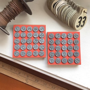 May include: Two red square wooden blocks with gray circular number stamps from 1 to 25 and 26 to 50. The numbers are in a vintage style. Other vintage items are in the background.