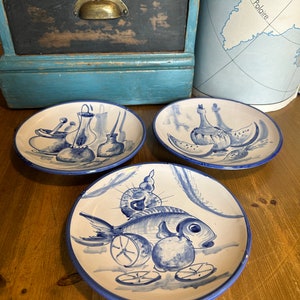May include: Three blue and white ceramic plates with hand-painted designs. The plates feature a fish, lemons, and a bottle of wine, a bottle of wine and watermelon, and a bottle of wine and a mortar and pestle.