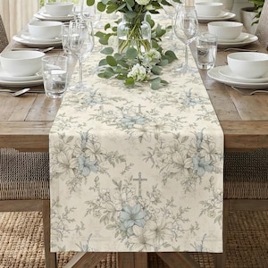 Peut inclure: Une table à manger en bois, ornée d'un chemin de table floral avec un motif de croix. Des chaises en osier entourent la table, avec des assiettes, des bols et des verres blancs pour chaque couvert. Un vase de fleurs est au centre.