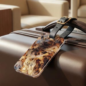 May include: A brown suitcase with a leather handle and a luggage tag. The luggage tag has a tortoiseshell pattern and is attached to the handle with a black leather strap and a gold buckle. The suitcase is in a well-lit room.
