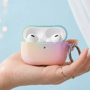 May include: A protective case for wireless earbuds featuring a smooth gradient finish that transitions from soft blue to pink. The case is open, revealing two white earbuds inside, and includes a circular metal ring attachment on the side.