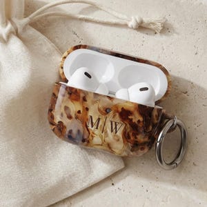 May include: An open AirPod case with a tortoiseshell design, silver ring, and the initials "MW" is next to a drawstring pouch. The case holds white AirPods. The pouch and background are a neutral beige color.