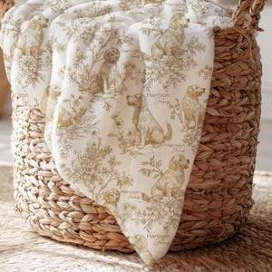 May include: A white blanket featuring a repeating toile-style pattern in a tan color, depicting golden retriever dogs sitting among floral arrangements and foliage. The text Harrison James is printed in a script font next to each dog illustration.