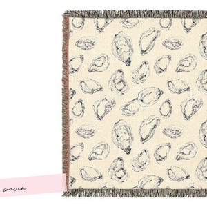 May include: Cream-coloured woven throw blanket featuring a pattern of black oyster illustrations. The blanket has a fringed edge. The word "woven" is written on a pink label.
