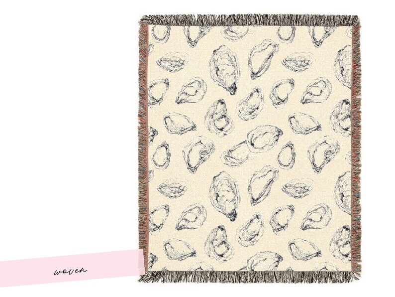 May include: Cream-coloured woven throw blanket featuring a pattern of black oyster illustrations. The blanket has a fringed edge. The word "woven" is written on a pink label.