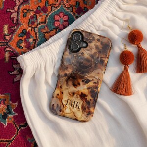 May include: A brown and black tortoiseshell patterned phone case with the letters JMK printed in black, accompanied by a pair of dangling earrings featuring orange pom-poms and matching orange silk tassels with gold-toned metal hooks.