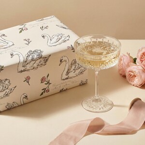 Close view of personalized swan bridal wrapping paper beside a coupe glass blush roses and silk ribbon showing elegant wedding gift packaging with soft romantic styling