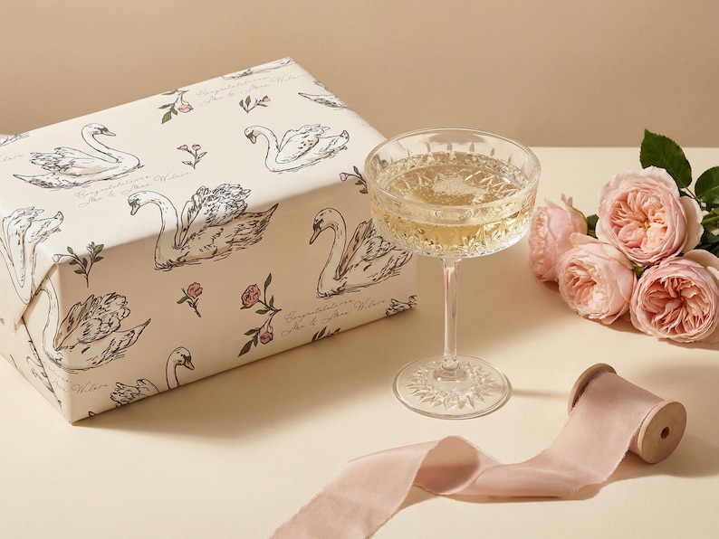 Close view of personalized swan bridal wrapping paper beside a coupe glass blush roses and silk ribbon showing elegant wedding gift packaging with soft romantic styling