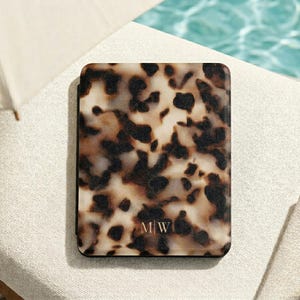 May include: A rectangular e-reader case featuring a brown, black, and cream tortoiseshell pattern. The bottom centre of the case displays the monogram letters M|W in a thin, light-coloured font. The case has a smooth, flat, and solid surface.