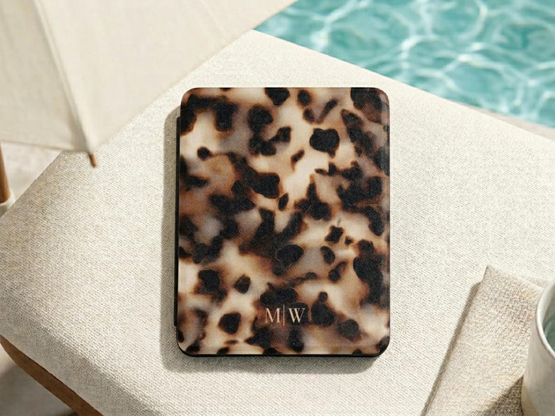 May include: A rectangular e-reader case featuring a brown, black, and cream tortoiseshell pattern. The bottom centre of the case displays the monogram letters M|W in a thin, light-coloured font. The case has a smooth, flat, and solid surface.