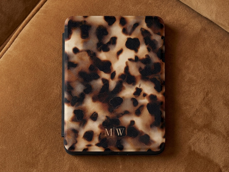 May include: A rectangular e-reader case featuring a brown, black and cream tortoiseshell pattern. The bottom centre of the case displays the gold-coloured initials M|W in a serif font. The case has a smooth finish and rounded corners.