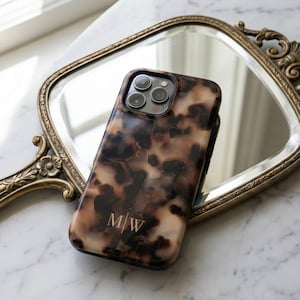 May include: A smartphone case featuring a brown and cream tortoiseshell pattern with a glossy finish. The case includes a triple-lens camera cutout at the top and the gold-coloured monogram letters M/W printed in the lower centre of the back panel.