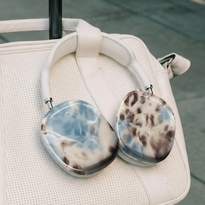 以下が含まれることがあります： 白のオーバーイヤーヘッドホン。青、茶色、白の色合いの大理石模様のデザインで、白い革のバッグの上に置かれています。バッグには穴あき模様とハンドルが付いています。ヘッドホンは白いヘッドバンドとシルバーのアクセントが付いています。