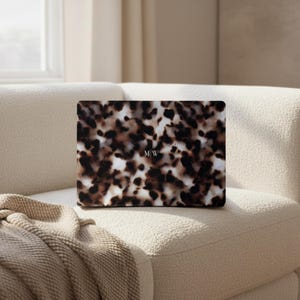 May include: A rectangular laptop skin featuring a brown, black, and cream tortoiseshell pattern. The center of the skin displays the letters M|W in a small, white, serif font. The design covers the entire surface of the laptop lid.