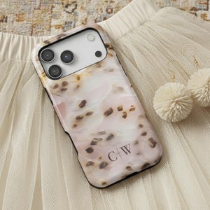 May include: A smartphone case with a light pink and white marbled pattern featuring dark brown spots and the initials C|W printed in black. Beside the case are two cream-coloured yarn pom-pom earrings with gold-toned metal hooks.