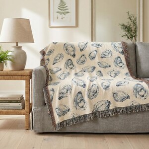 May include: A cream-coloured throw blanket with a repeating blue oyster shell pattern is draped over a grey sofa. The blanket has a fringed edge. A lamp and side table are to the left.