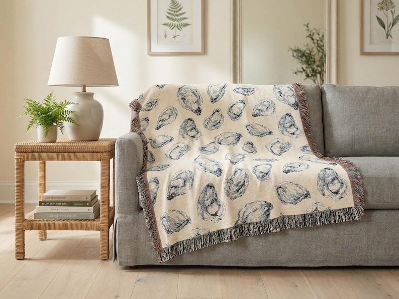 May include: A cream-coloured throw blanket with a repeating blue oyster shell pattern is draped over a grey sofa. The blanket has a fringed edge. A lamp and side table are to the left.