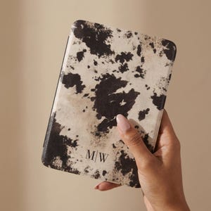 May include: A rectangular e-reader case featuring a black and cream cow print pattern. The bottom center of the case displays the black serif letters M|W. The case has a textured surface and rounded corners, providing a protective cover for a device.