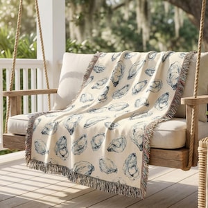 May include: A cream-coloured throw blanket with a blue oyster shell pattern draped over a wooden swing seat. The blanket has a fringed edge and is on a porch with a wicker side table and a mug.